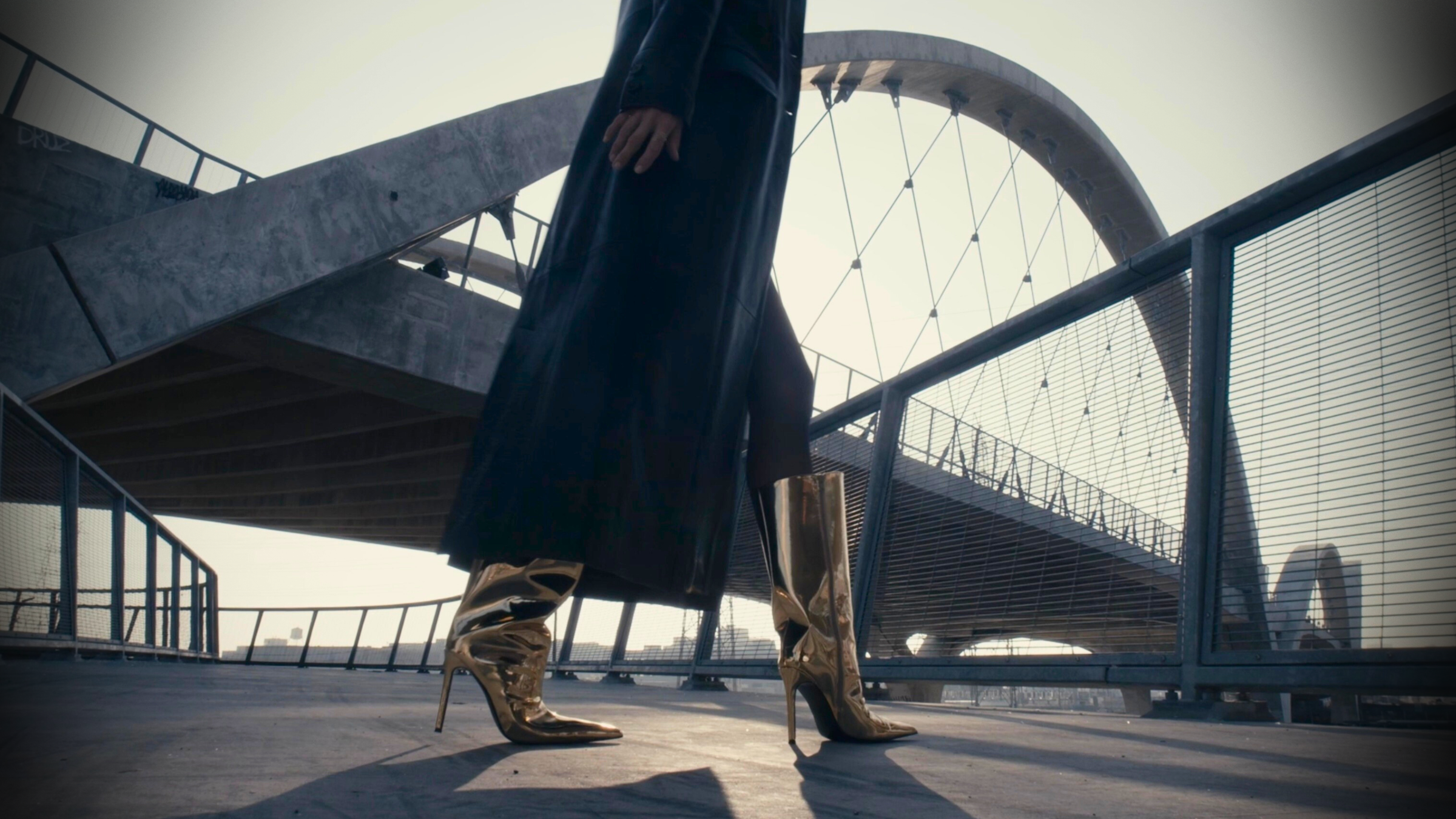 Person wearing a long coat and high-heeled shoes standing on a bridge.